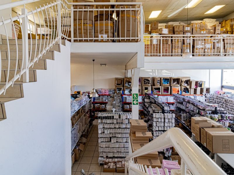 Commercial warehouse space featuring a mezzanine level, metal staircase, and tiled flooring, currently utilized for storage and inventory management.