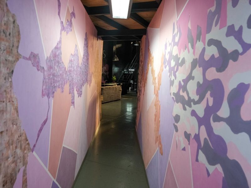 A narrow industrial hallway featuring exposed ceiling beams, concrete flooring, and decorative wall murals, leading towards an open-plan area.