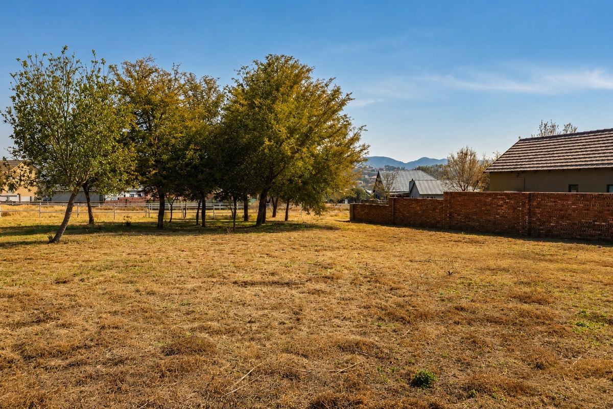 A spacious vacant residential plot featuring mature trees, a brick boundary wall, and clear views of the surrounding landscape.