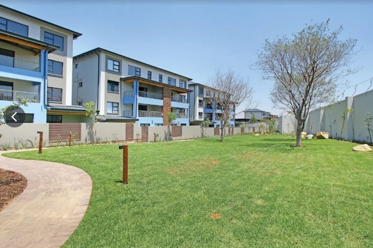 A well-maintained communal garden area featuring a paved walkway, manicured lawn, and boundary wall, set against the backdrop of modern apartment buildings.