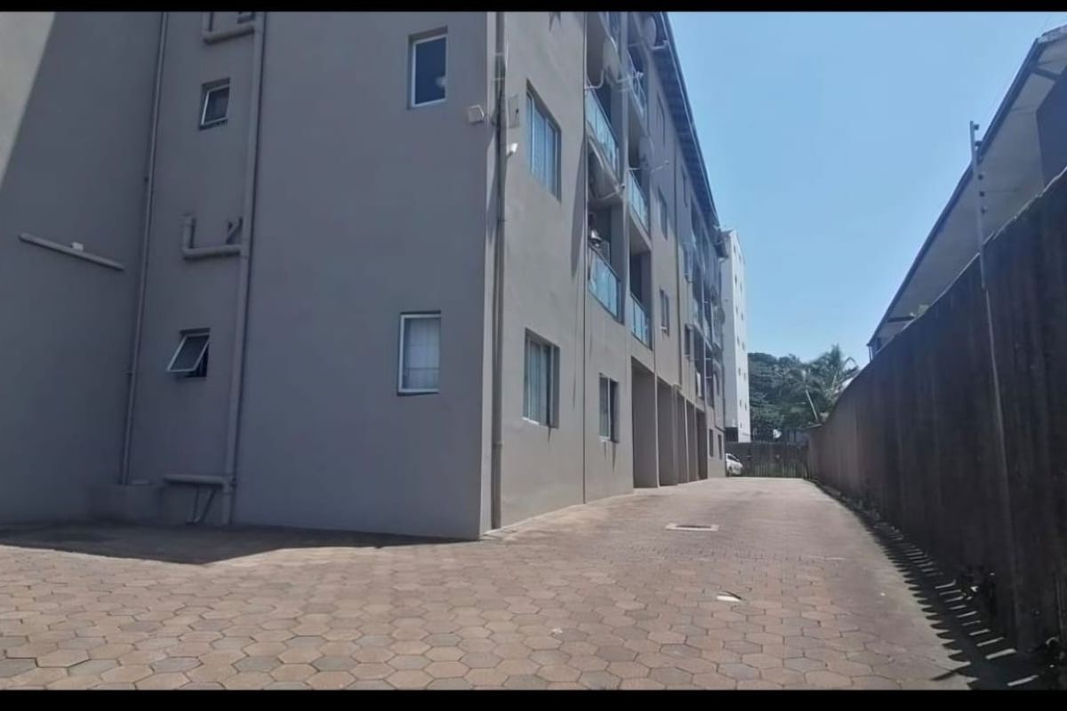 Exterior view of the apartment building showing the paved driveway, side facade, and boundary wall with security fencing.