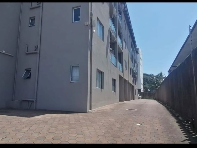 Exterior view of the apartment building showing the paved driveway, side facade, and boundary wall with security fencing.