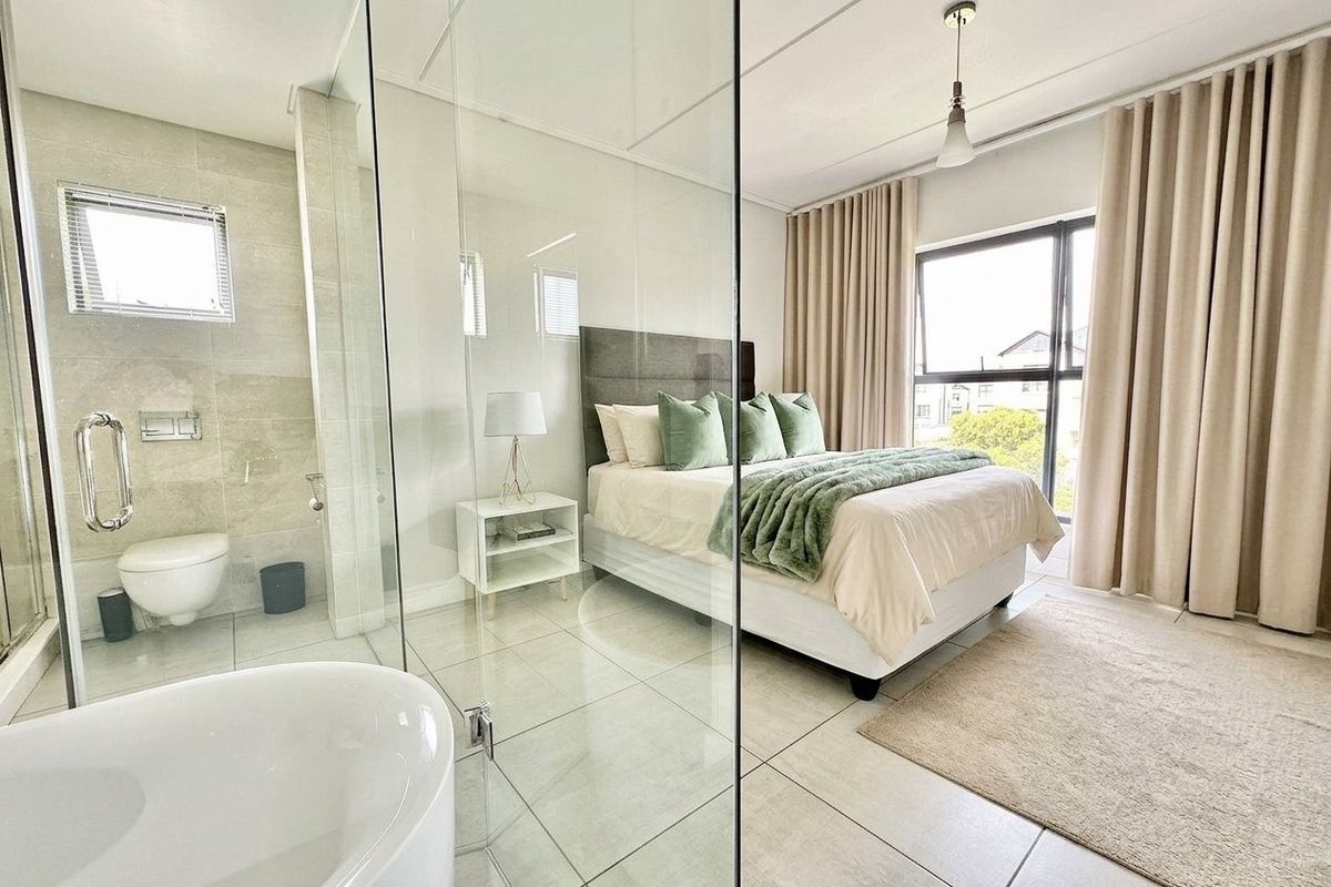 Modern bedroom featuring an open-plan glass-enclosed en-suite bathroom with a freestanding tub and wall-mounted toilet, complemented by large windows and neutral finishes.
