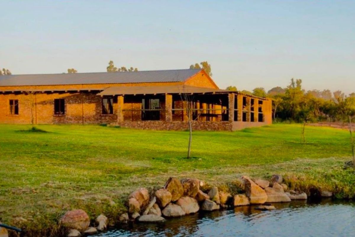 A rustic stone-built farmhouse featuring a spacious covered patio, overlooking a tranquil pond and expansive green lawns.