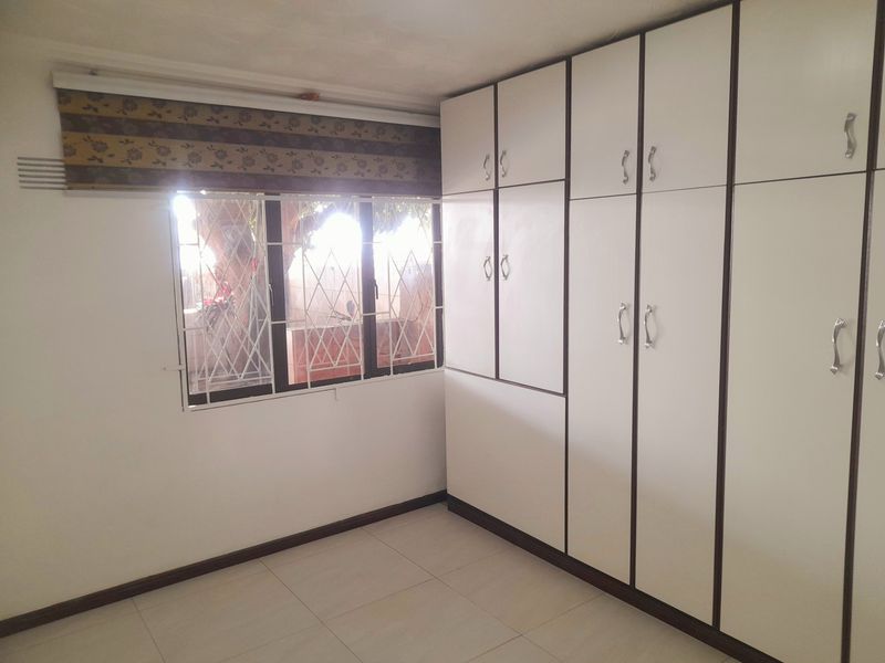 A clean, empty bedroom featuring extensive built-in white cupboards, tiled flooring, and a window fitted with security bars.