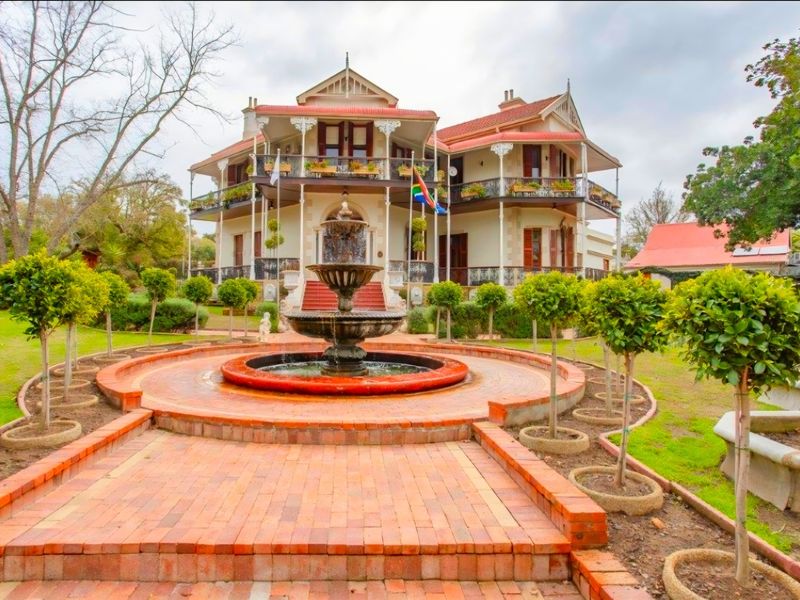 Grand historic residence featuring a prominent central fountain, manicured garden, and expansive wrap-around verandas with decorative wrought iron detailing.