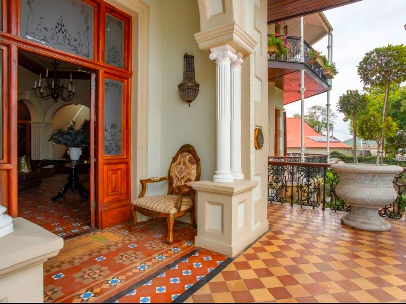 A beautifully maintained covered balcony featuring intricate decorative tiling, classical columns, and elegant wrought iron railings, providing a charming transition to the interior.