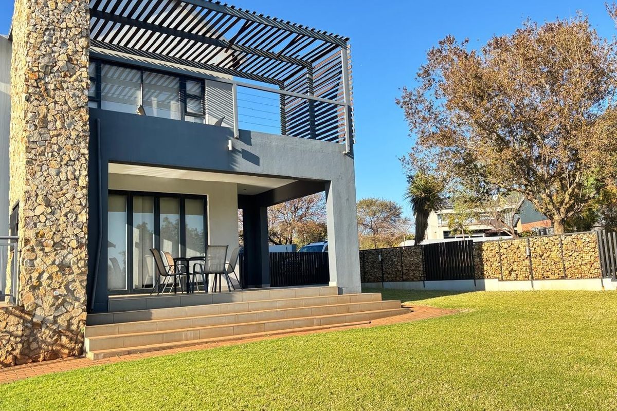 Rear view of a modern double-story house featuring a stone-clad chimney, a covered patio with tiled steps, and an upper-level balcony with a timber pergola.