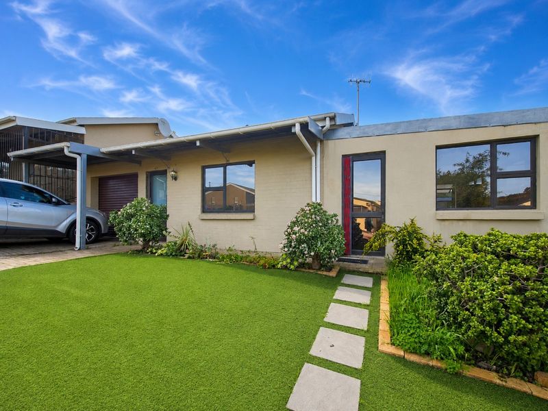 Front elevation of a single-story house featuring a carport, paved walkway, artificial lawn, and aluminium window frames under a clear blue sky.