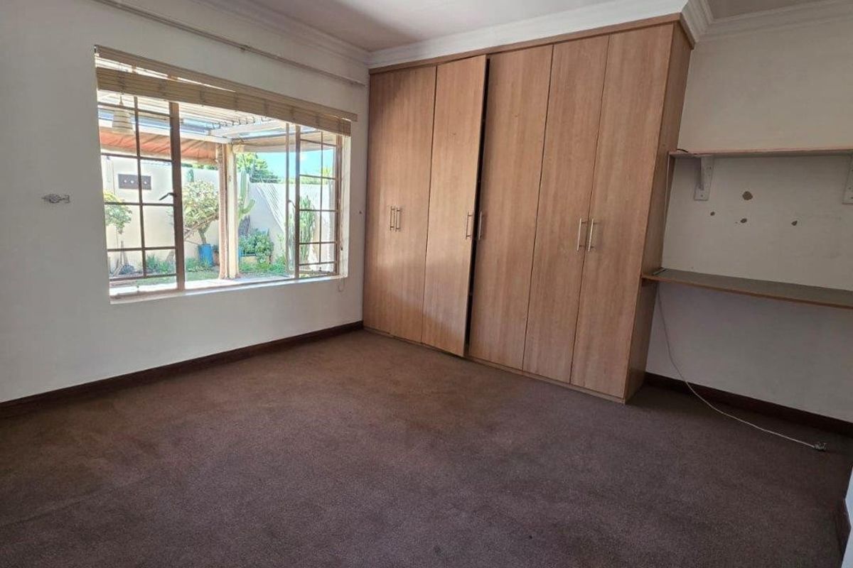 A vacant bedroom featuring built-in wooden cupboards, a built-in desk, carpeted flooring, and a window overlooking the garden area.