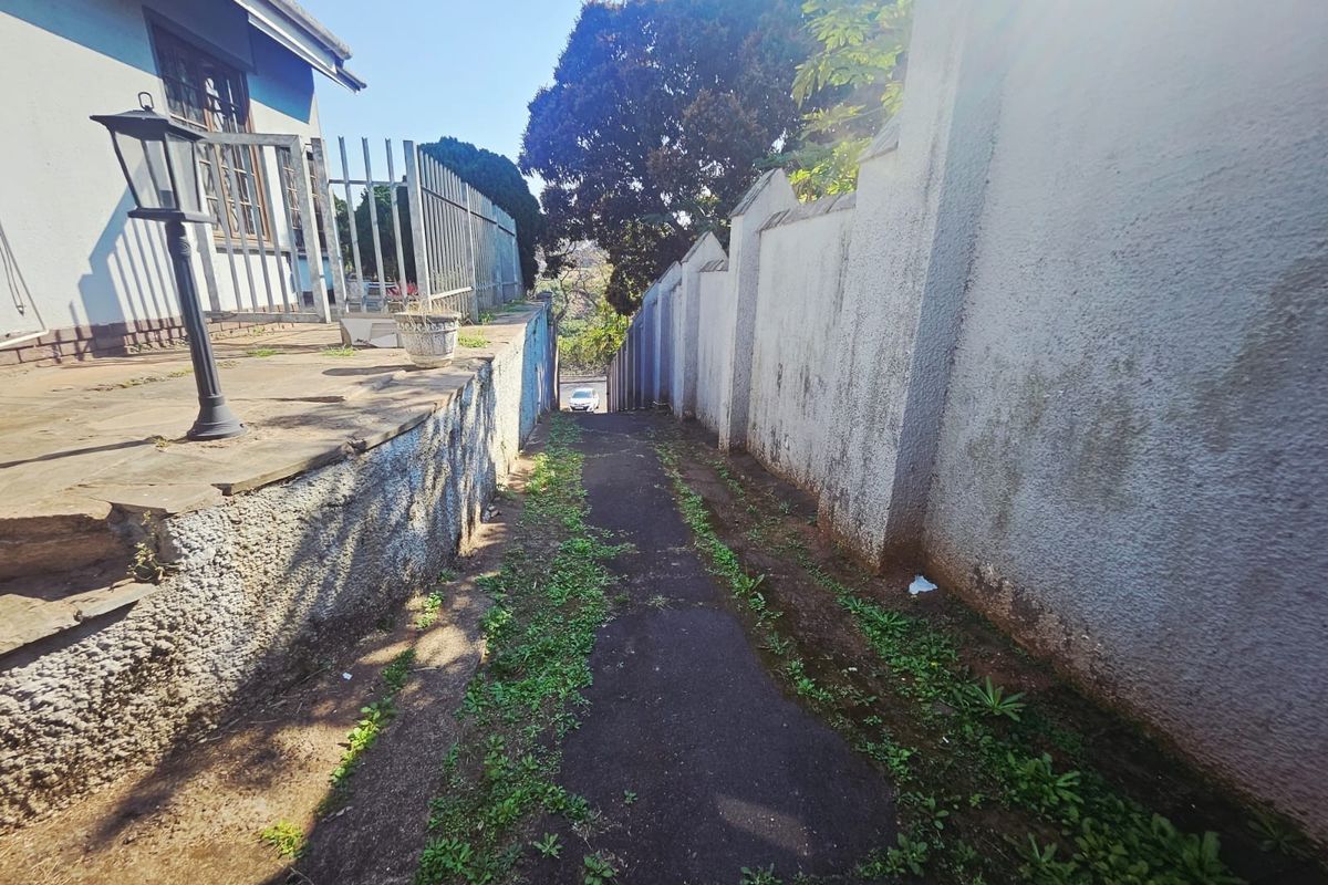 A narrow exterior side pathway featuring a paved walkway, boundary walls, and metal security fencing, showing signs of weathering and vegetation growth.