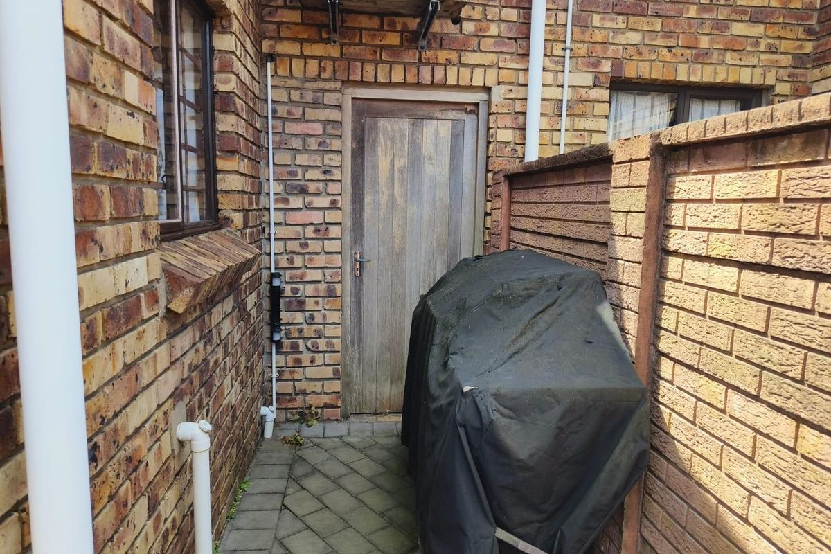 A narrow rear exterior passage featuring exposed brick walls, a paved walkway, and a weathered timber access door with an external air conditioning unit.