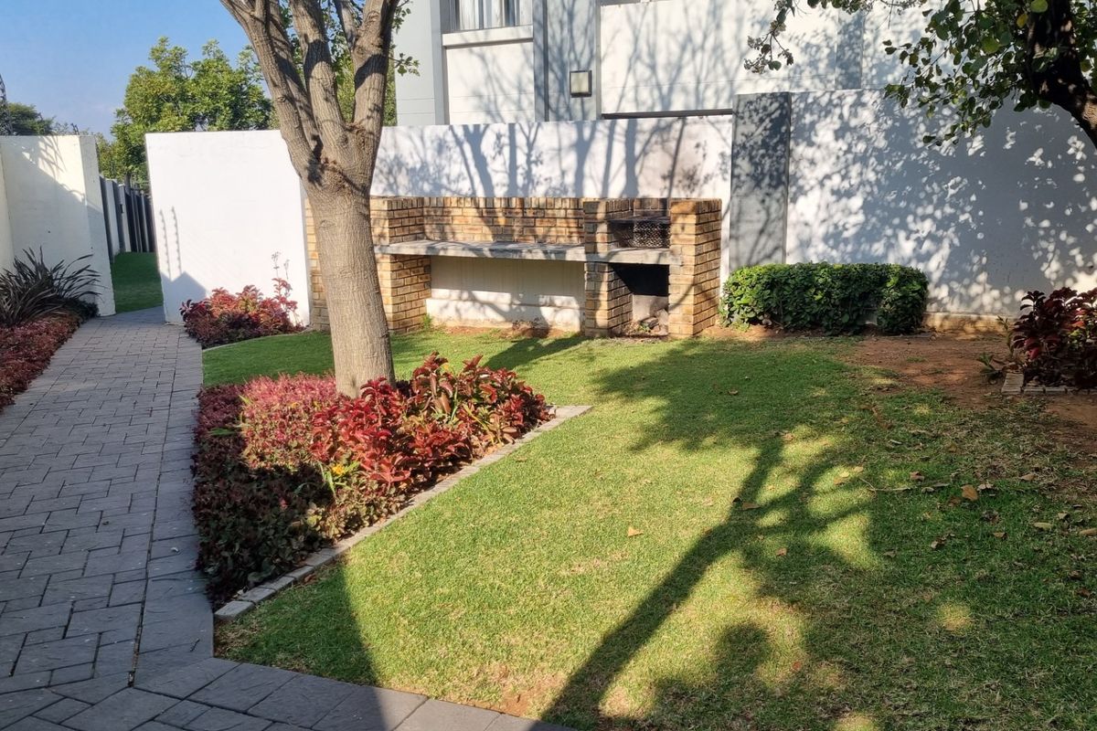 A well-maintained rear garden featuring a paved walkway, established trees, and a built-in brick braai area against the boundary wall.