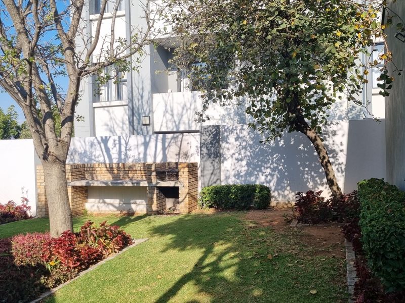A private rear garden area featuring a built-in brick braai, paved walkway, and established landscaping adjacent to the apartment building.