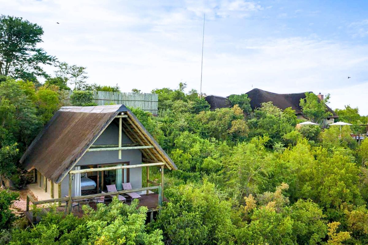 Elevated view of a rustic thatch-roofed cabin nestled in lush greenery, featuring a private timber deck and sliding glass doors.