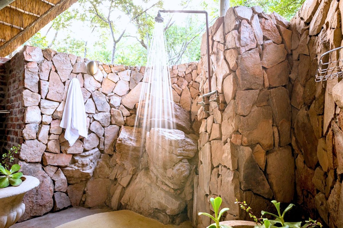 An outdoor shower area featuring natural stone wall cladding, exposed plumbing fixtures, and a rustic thatched roof covering.