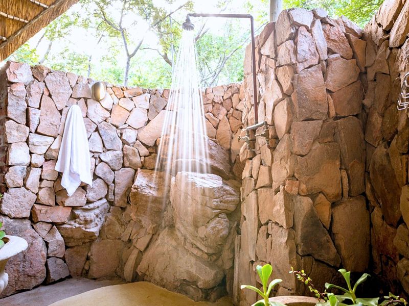 An outdoor shower area featuring natural stone wall cladding, exposed plumbing fixtures, and a rustic thatched roof covering.