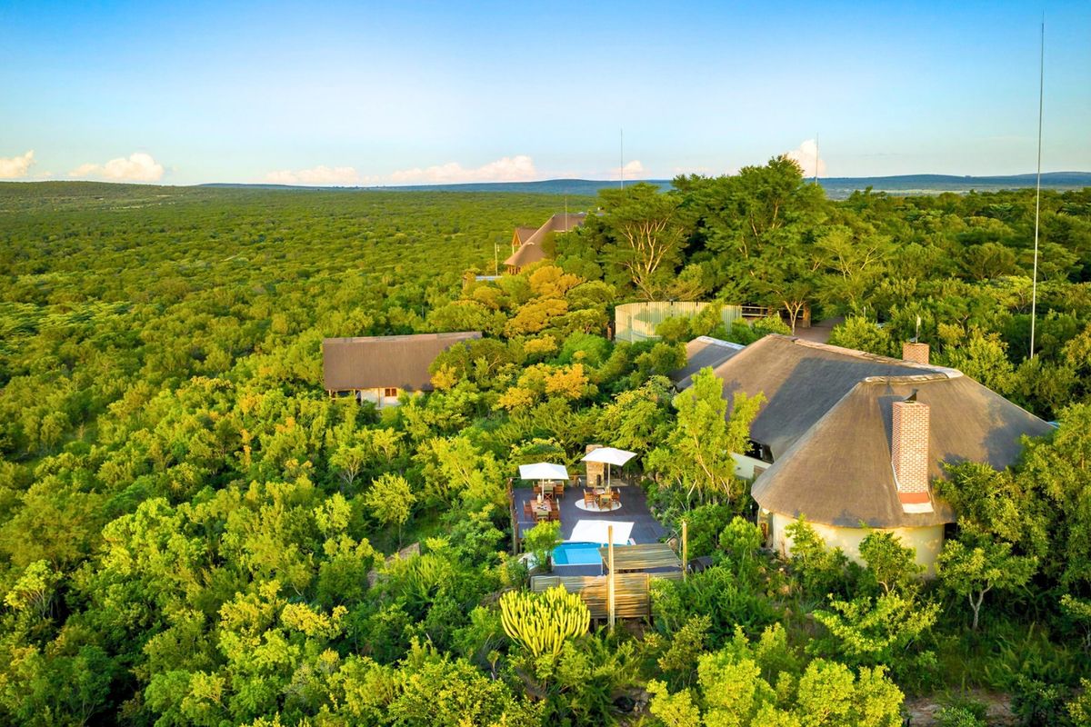 Aerial view of a luxury farm property featuring thatched-roof buildings, a swimming pool, and a timber deck nestled within a lush natural landscape.