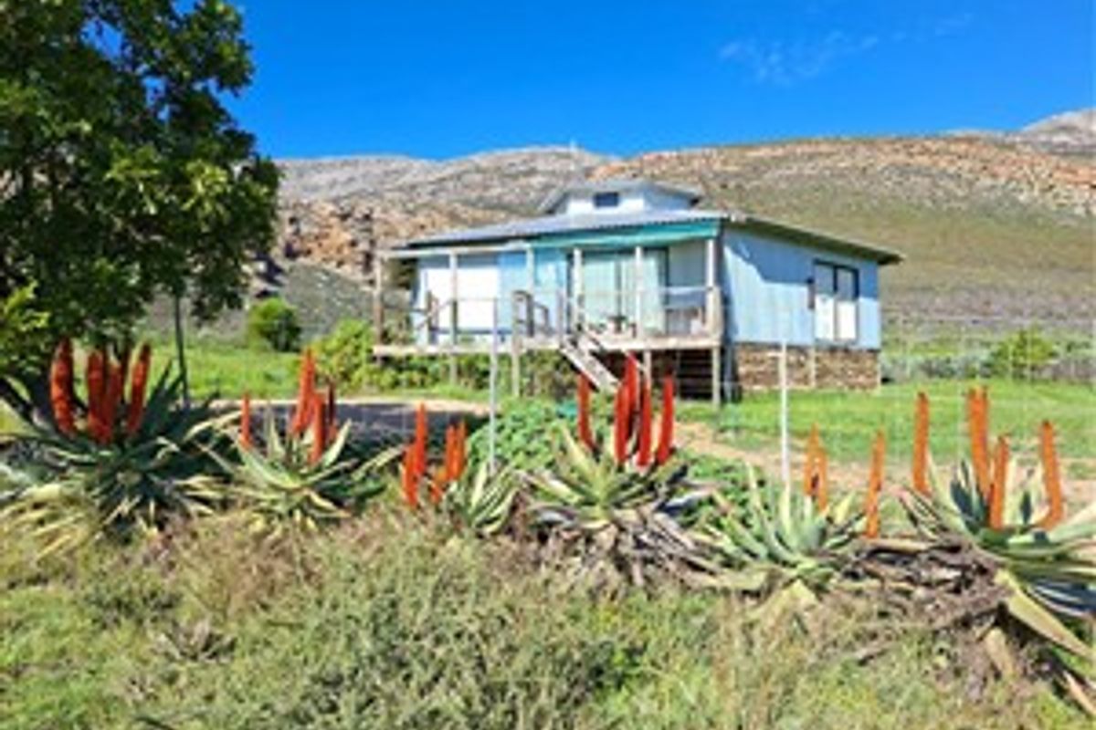 A rural farm cottage featuring a raised timber deck, metal roofing, and sliding glass doors, set against a scenic mountain backdrop.