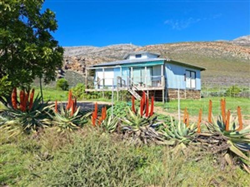 A rural farm cottage featuring a raised timber deck, metal roofing, and sliding glass doors, set against a scenic mountain backdrop.