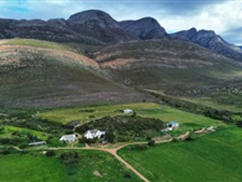 Aerial view of a farm property featuring multiple buildings, surrounding green pastures, and a dirt access road set against a mountain backdrop.