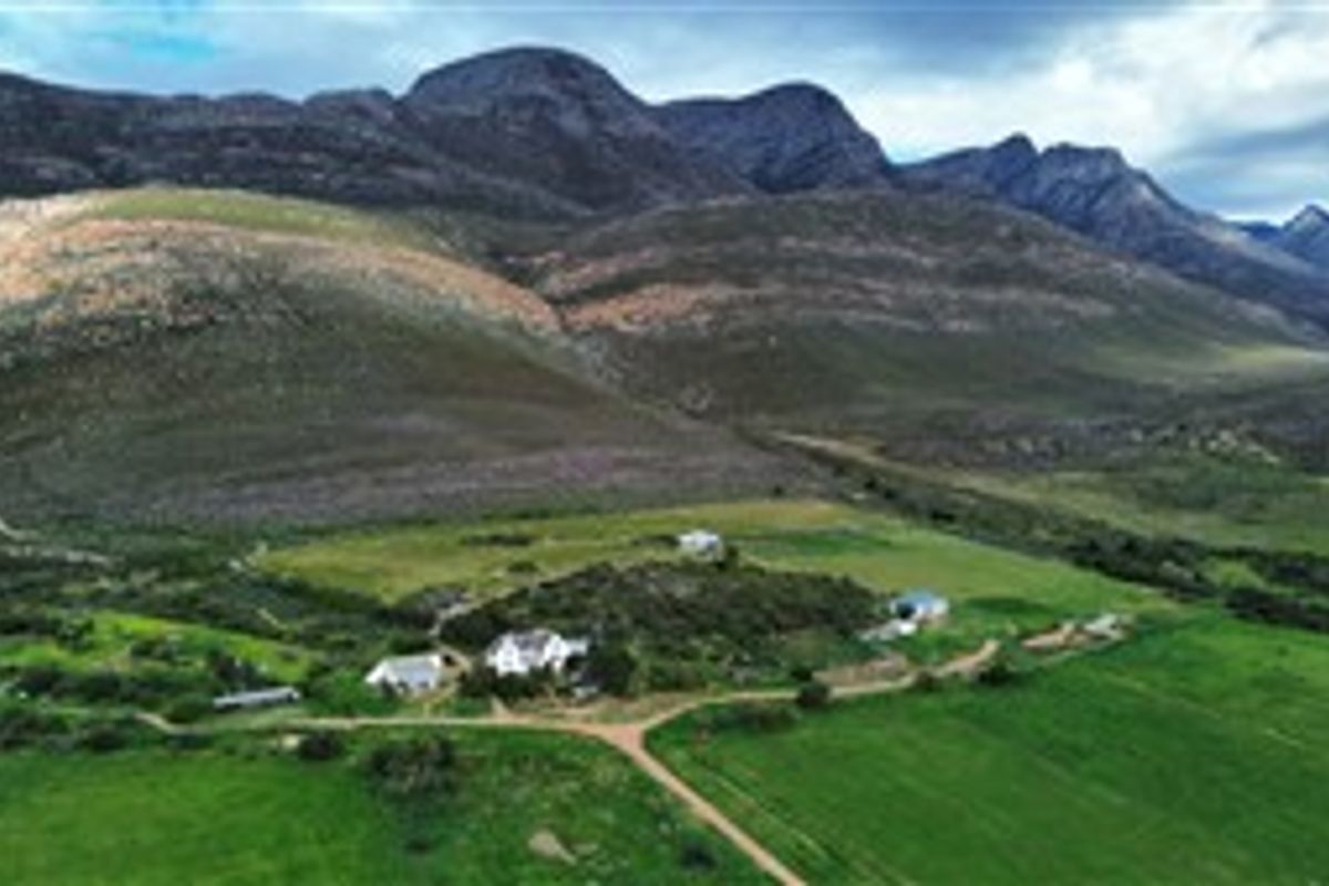 Aerial view of a farm property featuring multiple buildings, surrounding green pastures, and a dirt access road set against a mountain backdrop.