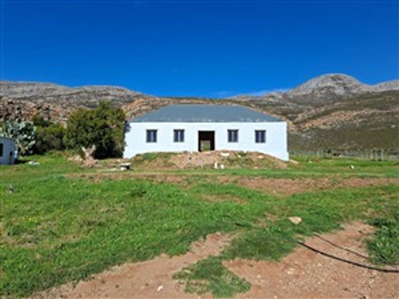 A front view of a rural farmhouse featuring a metal roof and mountain backdrop, set on an undeveloped plot of land.