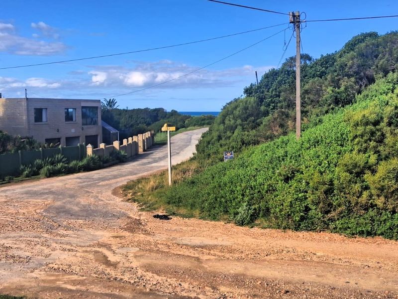 A view of the unpaved access road leading towards the coast, featuring surrounding vegetation, utility infrastructure, and nearby residential boundary walls.