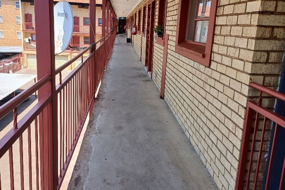 An exterior apartment walkway featuring a brick facade, metal safety railings, and a concrete floor, providing access to multiple residential units.