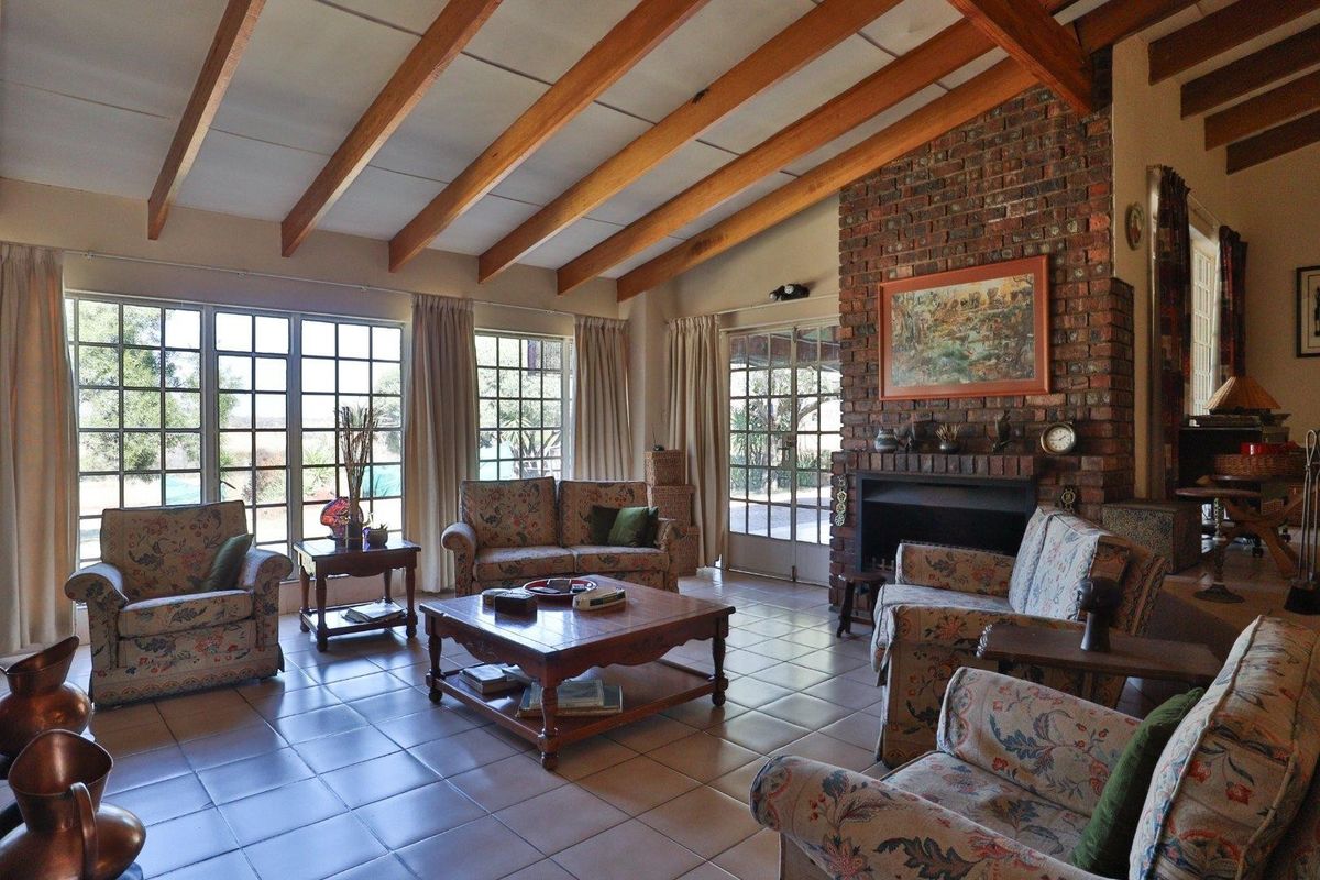Spacious living room featuring exposed timber ceiling beams, a classic brick fireplace, tiled flooring, and large windows providing ample natural light.