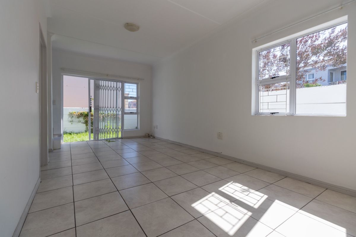 A bright, empty living room featuring neutral tiled flooring, large windows, and a sliding door with a security gate leading to the exterior.