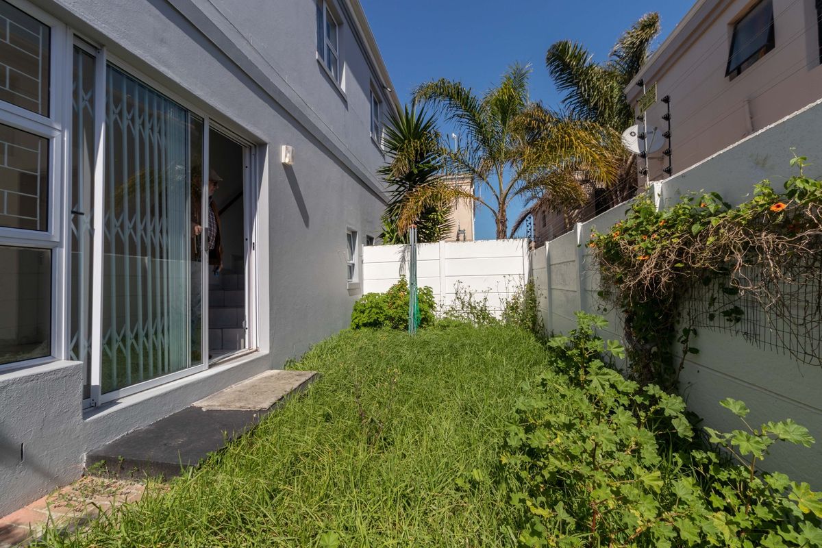 A rear exterior view showing a small garden area with overgrown grass, enclosed by a precast concrete wall with electric fencing and a sliding door.