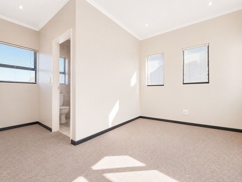 A bright, neutral-toned bedroom featuring carpeted flooring, recessed lighting, and an adjoining en-suite bathroom, offering a clean and versatile living space.