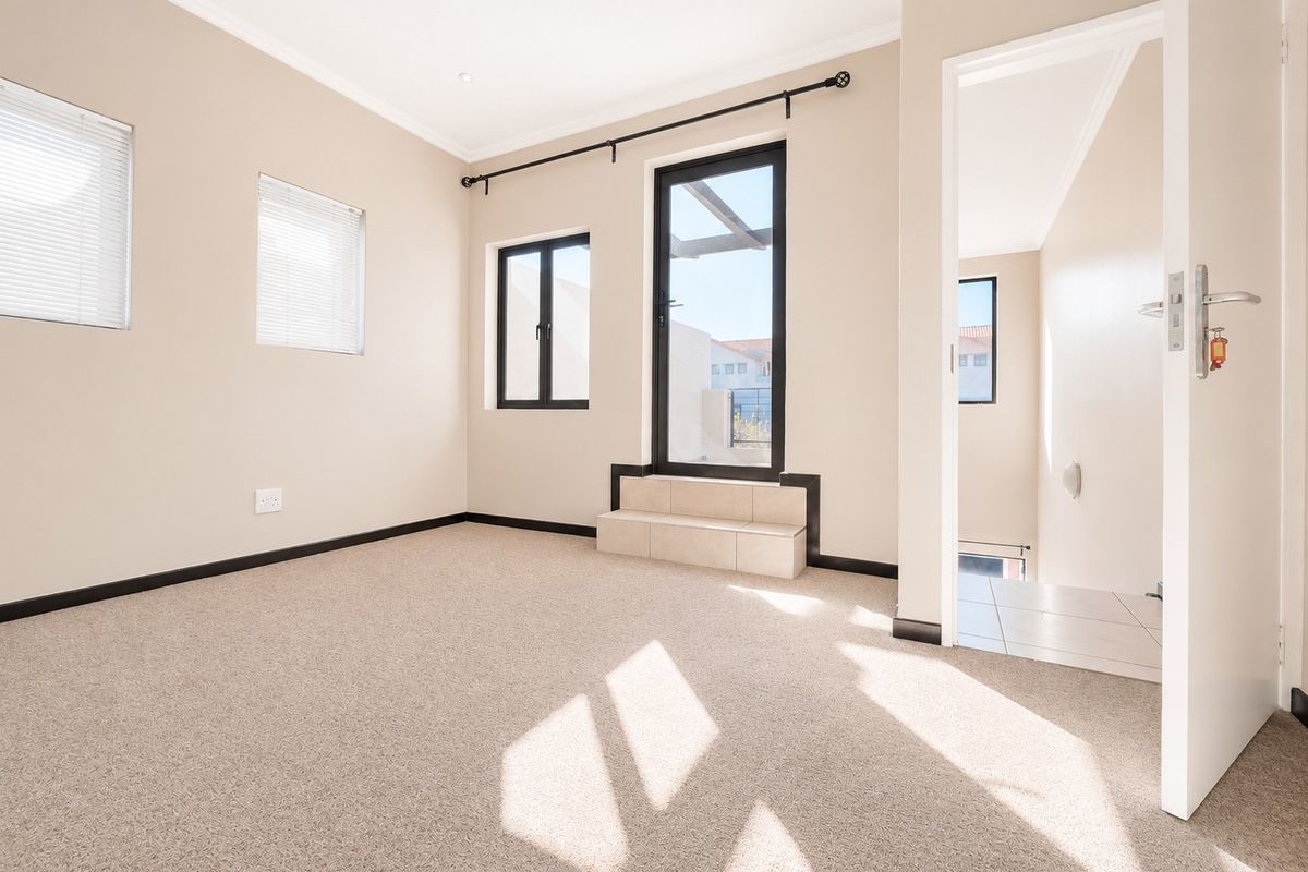 A bright, empty bedroom featuring neutral carpeting, aluminium window frames, and a tiled step leading to an exterior door.