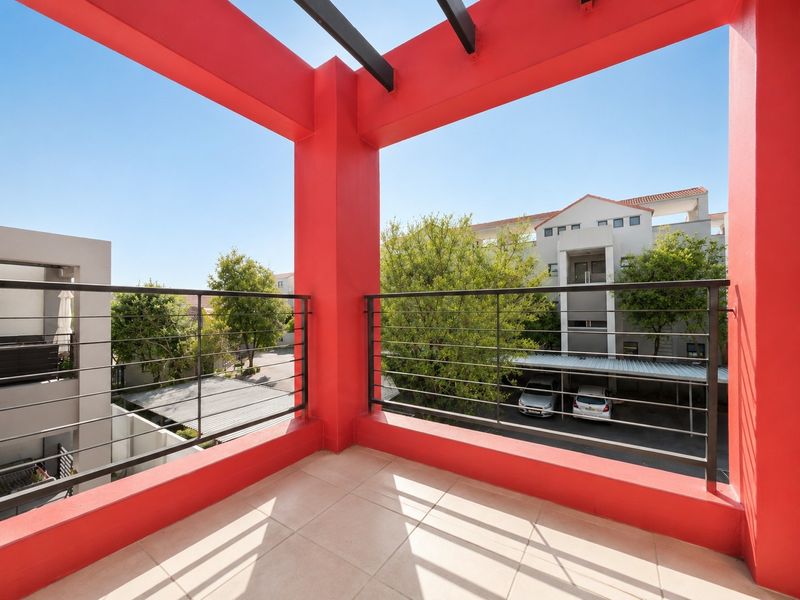 A bright, open-air balcony featuring tiled flooring, a metal balustrade, and a bold red pergola structure overlooking the complex parking area.