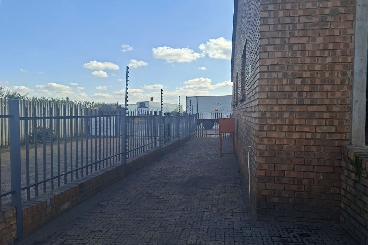 A paved side walkway along a brick industrial building, secured by steel palisade fencing with integrated electric security wires.