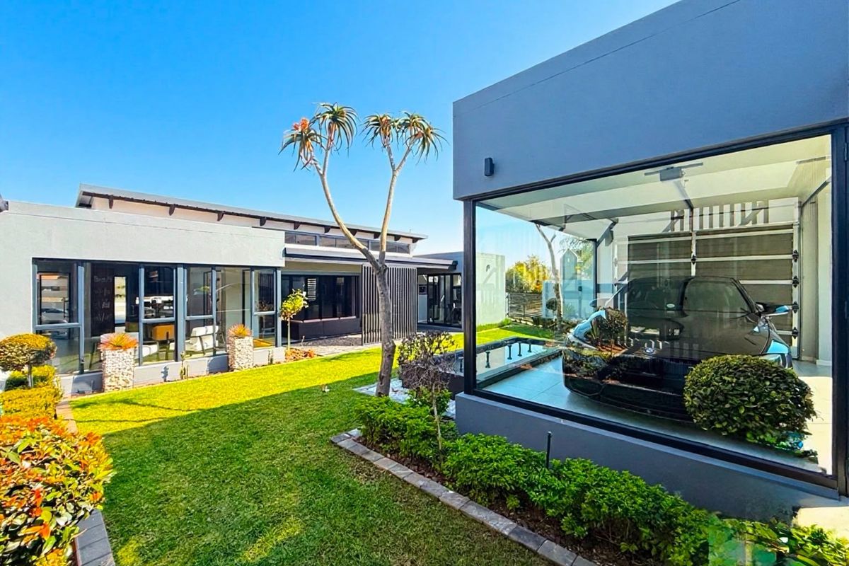 Modern architectural rear exterior featuring a glass-walled garage, manicured lawn, and a contemporary water feature in a well-maintained garden setting.