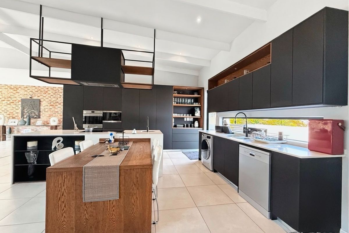 Modern kitchen featuring sleek dark cabinetry, a large wooden island, integrated appliances, and a striking brick feature wall under a vaulted ceiling.