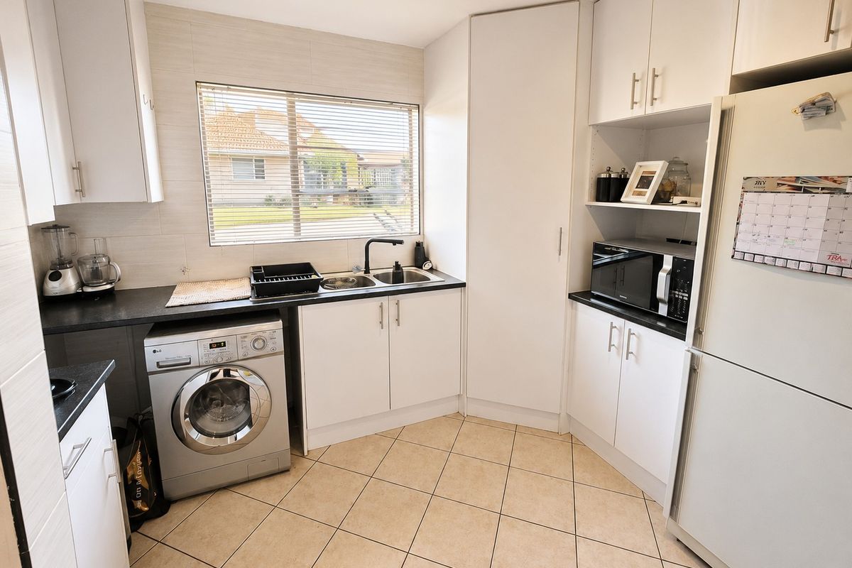 A functional kitchen featuring white cabinetry, dark countertops, tiled splashbacks, and integrated laundry plumbing, offering a clean and practical workspace.