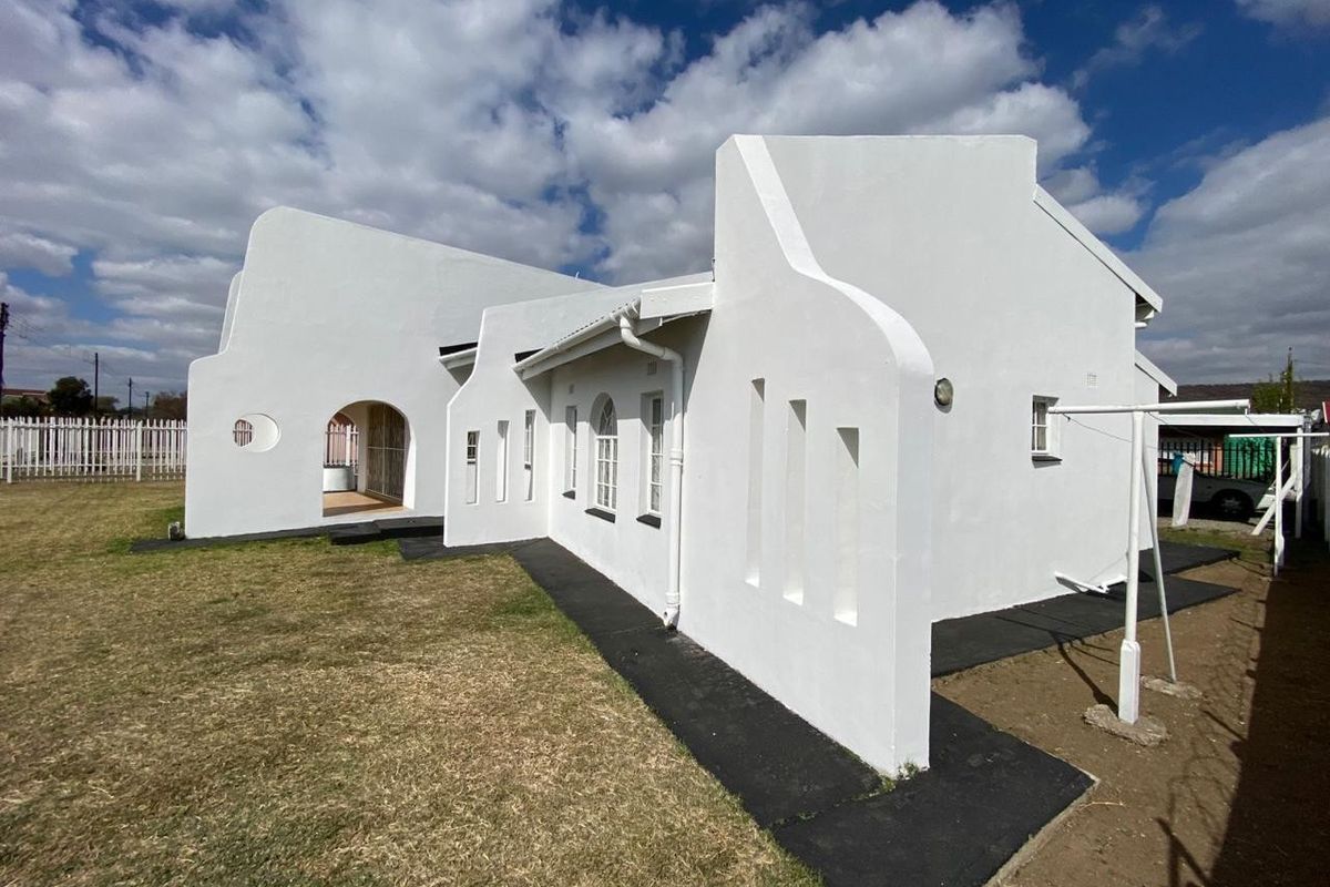 Rear view of a white house featuring distinctive gabled parapet walls, an arched entrance, and a paved walkway surrounding the structure.