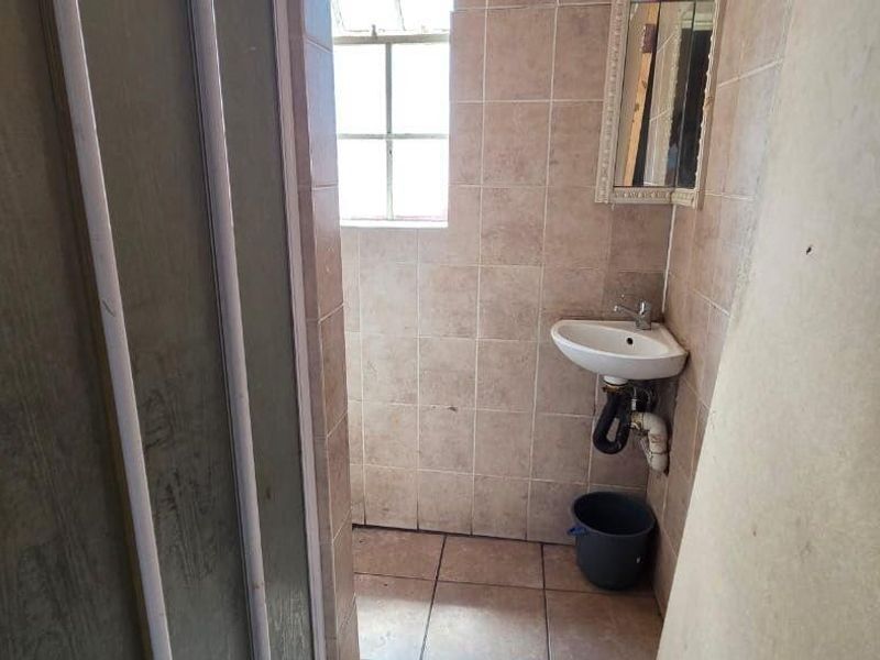 A small, dated bathroom featuring a wall-mounted basin, tiled surfaces, and a shower enclosure, currently in need of renovation and cosmetic updates.