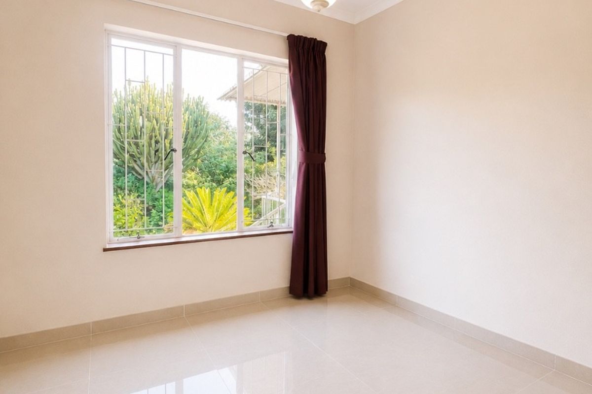 A bright, empty bedroom featuring neutral tiled flooring, a pendant light fixture, and a window with security bars overlooking the garden.