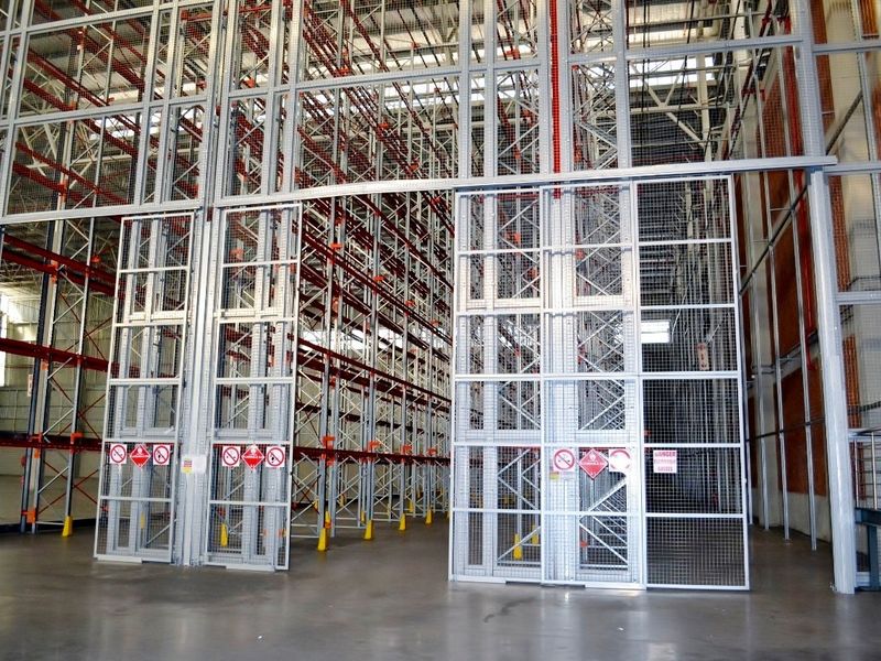 Large industrial warehouse space featuring extensive high-bay racking systems, secure metal mesh partitions, and sliding access gates with durable concrete flooring.
