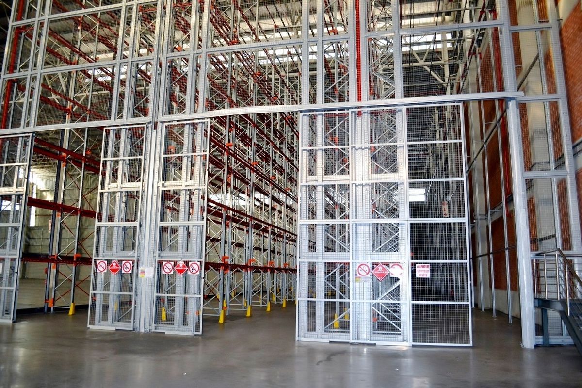 Large industrial warehouse space featuring extensive high-bay racking systems, secure metal mesh partitions, and sliding access gates with durable concrete flooring.
