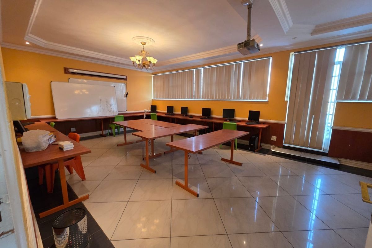 Spacious office or training room featuring tiled flooring, built-in desk surfaces, ceiling-mounted projector, and large windows with vertical blinds.