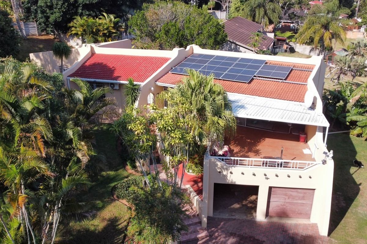 Aerial view of the property featuring a tiled roof with solar panels, a covered patio area, and a well-maintained garden with a garage.
