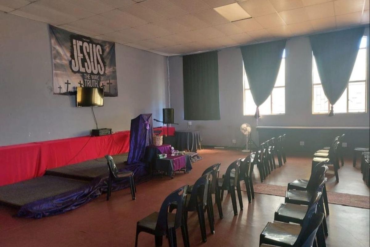A multi-purpose hall featuring a raised stage area, suspended ceiling, and large windows providing natural light, suitable for community or commercial gatherings.
