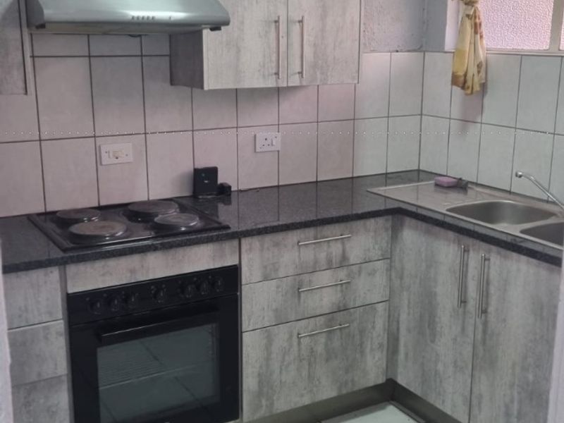 Compact kitchen featuring light grey wood-look cabinetry, dark countertops, built-in oven and hob, extractor fan, and tiled finishes throughout.