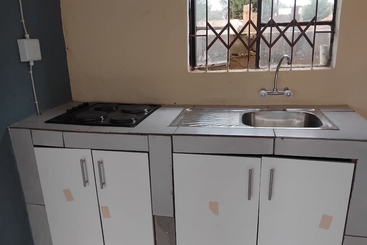 A compact kitchen area featuring a tiled countertop, built-in electric hob, stainless steel sink, and basic cabinetry with security bars on the window.