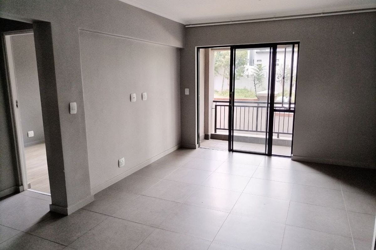 A bright, empty living room featuring neutral tiled flooring and a sliding glass door providing direct access to a private balcony.